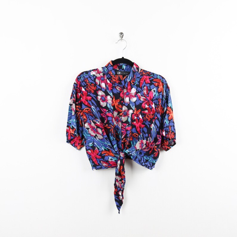 May include: A blue, red, and white floral print cropped shirt with a tie front. The shirt has short sleeves and a button-up collar.
