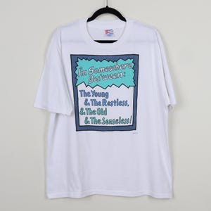 May include: White t-shirt with a blue and green graphic print that says "I'm Somewhere Between: The Young & The Restless, & The Old & The Senseless!"