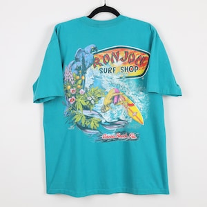 Puede incluir: Camiseta turquesa con un gr&aacute;fico de un loro azul, flores y un surfista en una tabla de surf amarilla. La camiseta tiene el texto "RON JON SURF SHOP" y "Cocoa Beach, Fla."