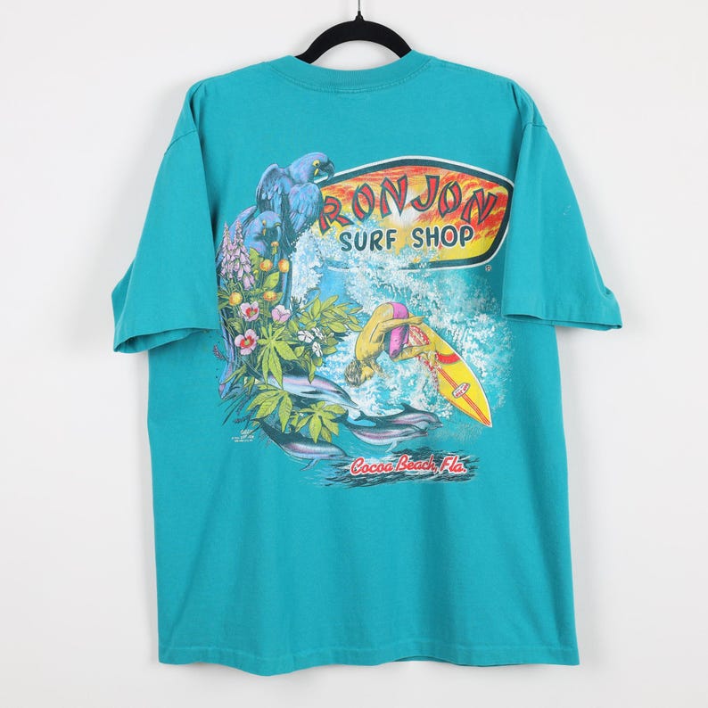 Puede incluir: Camiseta turquesa con un gr&aacute;fico de un loro azul, flores y un surfista en una tabla de surf amarilla. La camiseta tiene el texto "RON JON SURF SHOP" y "Cocoa Beach, Fla."