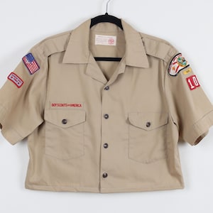 May include: Tan short-sleeved shirt with two front pockets and the words "BOY SCOUTS OF AMERICA" embroidered in red. Patches include an American flag and a patch with the number 10. The shirt is buttoned and hanging on a black hanger.