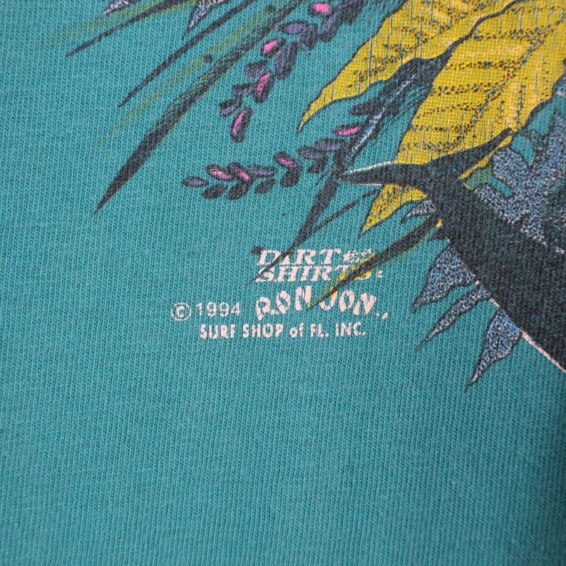 Puede incluir: Camiseta de color verde azulado con un dise&ntilde;o tropical con hojas amarillas y verdes y flores moradas. El texto "Darte Shirts" y "Ron Jon Surf Shop of FL. Inc." est&aacute;n impresos en la camiseta. La fecha de copyright es 1994.