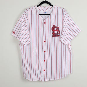 May include: White baseball jersey with red vertical stripes and red buttons. The jersey features the St. Louis Cardinals logo on the chest and a small logo on the sleeve. The jersey has short sleeves and a v-neck.