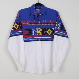 May include: A button-down shirt with a blue collar and yoke, and white sleeves. The shirt features a geometric pattern in red, yellow, black, and white across the chest and upper sleeves. The shirt is hanging on a black hanger.
