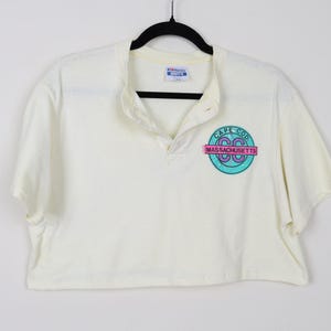 May include: Cream-colored cropped polo shirt with a button-up collar. The shirt features a circular graphic with the words "Cape Cod Massachusetts" in teal, pink, and white. The shirt is hanging on a black hanger.