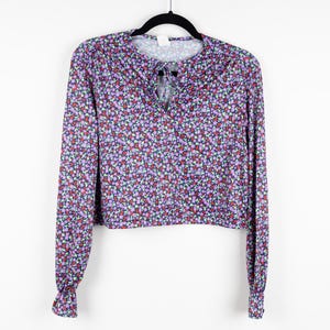 May include: A purple, red, and green floral print long-sleeve blouse with a tie at the neckline. The blouse has a cropped fit.