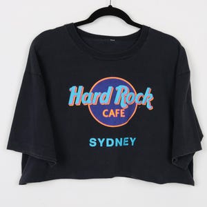 May include: Black crop top with the Hard Rock Cafe logo in blue, orange, and white, with the city name "SYDNEY" in blue below the logo. The shirt is hanging on a black hanger against a white background.