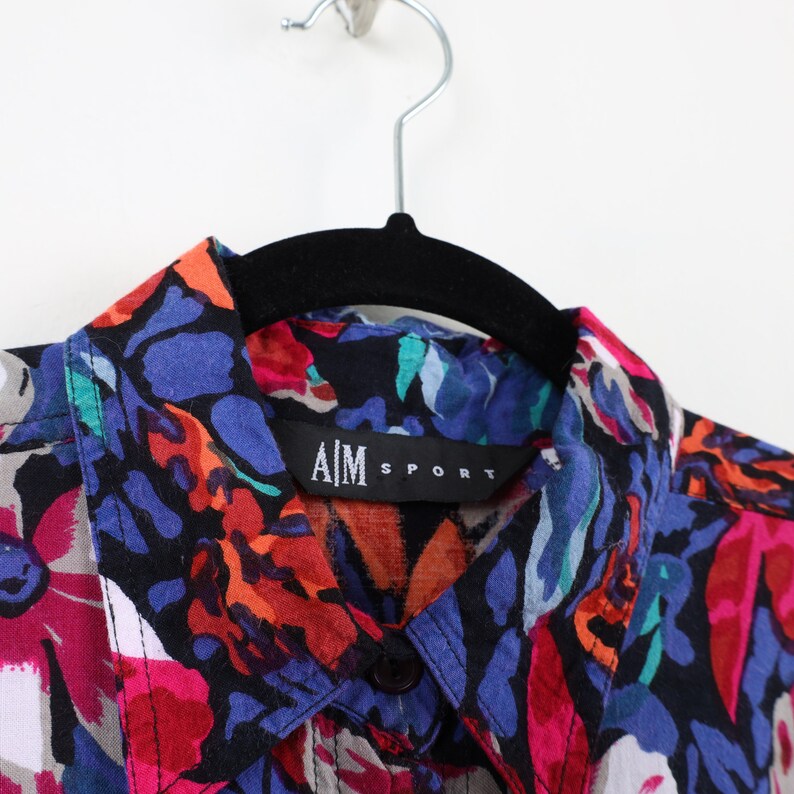 May include: A black button-up shirt with a colorful floral print. The shirt has a collar and a button closure. The label on the shirt reads "AIM SPORT".