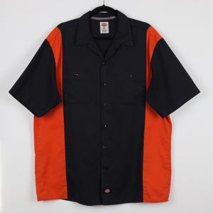 May include: A short-sleeve button-down shirt featuring a black center panel and collar with vibrant orange side panels. The shirt includes two chest pockets, a front button closure, and a Dickies brand label on the inner neck and bottom hem.