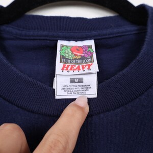May include: A navy blue t-shirt with a Fruit of the Loom "Heavy Cotton" tag. The tag is white with red and green grapes and an apple. The tag also says "100% Cotton Preshrunk, Assembled in El Salvador of U.S.A. Components, Care on Reverse".