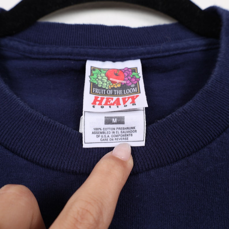 May include: A navy blue t-shirt with a Fruit of the Loom "Heavy Cotton" tag. The tag is white with red and green grapes and an apple. The tag also says "100% Cotton Preshrunk, Assembled in El Salvador of U.S.A. Components, Care on Reverse".