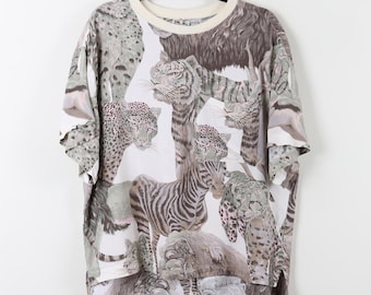 Könnte beinhalten: Weißes T-Shirt mit einem Aufdruck verschiedener Wildtiere, darunter Tiger, Zebras und Leoparden. Das T-Shirt hat einen cremefarbenen Rundhalsausschnitt und kurze Ärmel. Das Tiermusterdesign ist in Grau-, Braun- und Weißtönen gehalten.