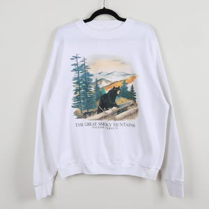 May include: White sweatshirt featuring a graphic of a black bear in a mountain landscape with pine trees. The text "THE GREAT SMOKY MOUNTAINS PIGEON FORGE TN" is printed below the graphic. The sweatshirt has long sleeves and a crew neck.