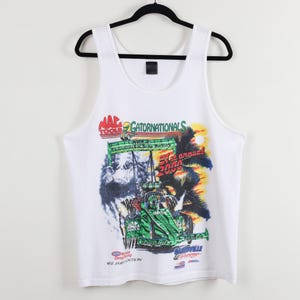 May include: White tank top with a colorful graphic featuring a race car, palm trees, and the text "Gatornationals 2000". The design includes the "Mac Tools" logo and other racing-related text. The tank top is hanging on a black hanger.