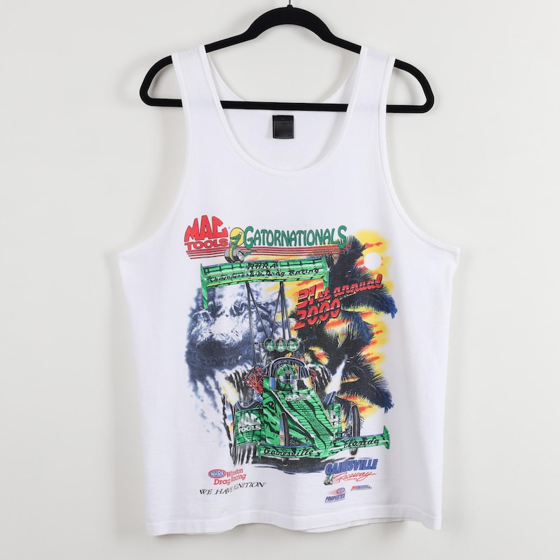 May include: White tank top with a colorful graphic featuring a race car, palm trees, and the text "Gatornationals 2000". The design includes the "Mac Tools" logo and other racing-related text. The tank top is hanging on a black hanger.