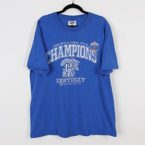 May include: A blue t-shirt with the text "1998 NCAA Final Four Champions" in white and gray. The shirt features the Kentucky Wildcats logo and the Lee brand label.