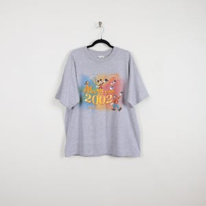 Pode incluir: Uma camiseta cinza com um gr&aacute;fico colorido de personagens da Disney, incluindo Mickey Mouse e Pateta. O design inclui o texto "EARS TO YOU" e "2002". A camiseta tem mangas curtas e gola redonda.