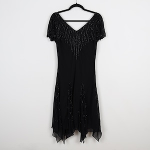 May include: A black dress with a V-neck and short sleeves, hanging on a black hanger. The dress features beaded embellishments along the neckline and down the front, with a flowing, asymmetrical hemline. The dress is made of a lightweight fabric.
