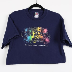 May include: A navy blue t-shirt with a rainbow-colored graphic of fireworks and the text "Girl Scouts Celebrating 90 Years! Girl Scouts of Santa Clara County".