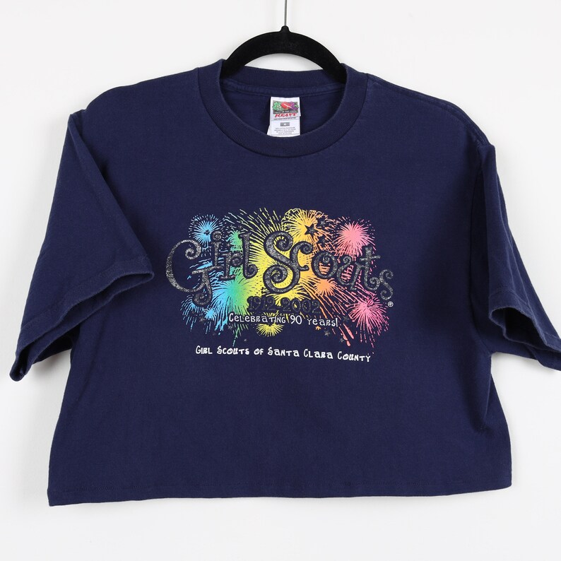 May include: A navy blue t-shirt with a rainbow-colored graphic of fireworks and the text "Girl Scouts Celebrating 90 Years! Girl Scouts of Santa Clara County".