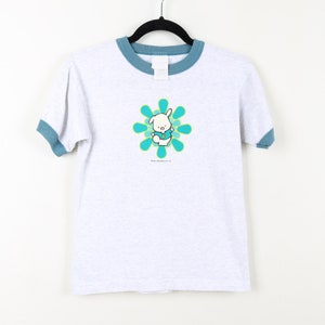 Puede incluir: Una camiseta blanca con cuello azul y un gr&aacute;fico de conejito de dibujos animados azul y verde. El conejito lleva una camisa azul con una letra "P" verde. El conejito est&aacute; de pie en una flor azul y verde.