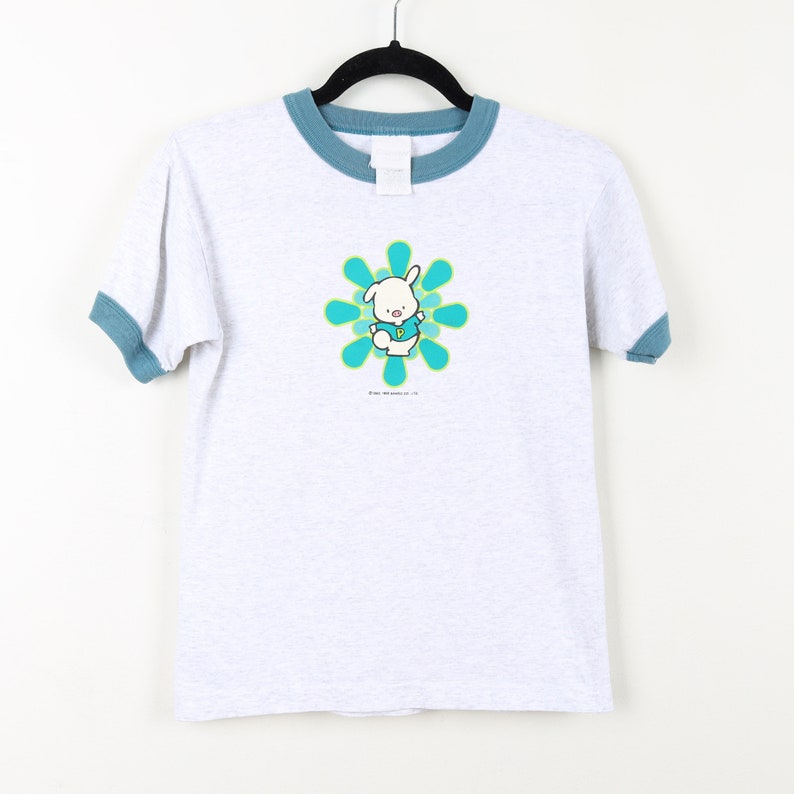 Puede incluir: Una camiseta blanca con cuello azul y un gr&aacute;fico de conejito de dibujos animados azul y verde. El conejito lleva una camisa azul con una letra "P" verde. El conejito est&aacute; de pie en una flor azul y verde.