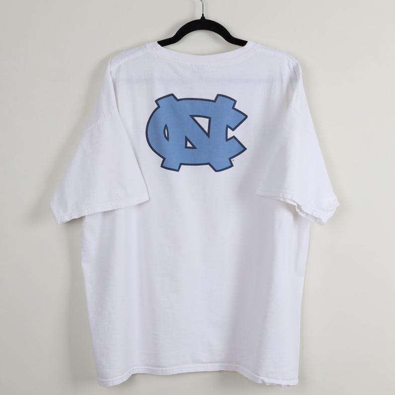 May include: White t-shirt with a light blue logo on the back. The logo is a stylized interlocking design. The shirt is hanging on a black hanger against a plain background. The shirt has short sleeves.