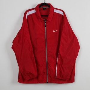 May include: A red Nike windbreaker jacket featuring a full-length front zipper with a black pull tab, a classic collar, and white stripes running along the top of the shoulders and sleeves. A small white Nike swoosh logo is embroidered on the chest.