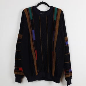 May include: Black sweater with a crew neck and long sleeves. The sweater features a geometric design with brown, green, red, and blue accents. The sleeves and shoulders have textured details. The sweater is hanging on a black hanger.