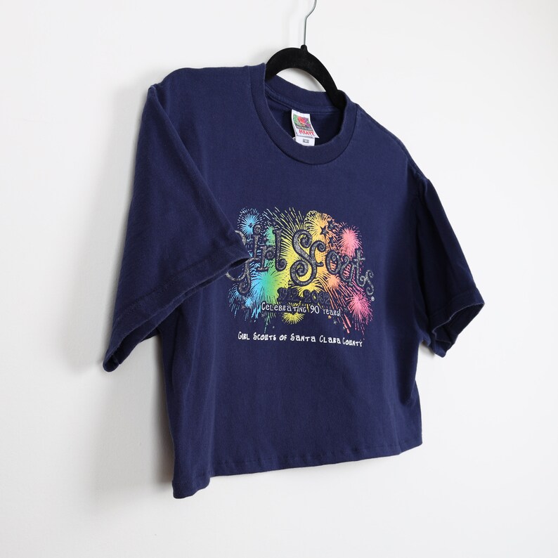 May include: A navy blue cropped t-shirt with a rainbow graphic of fireworks and the text "Girl Scouts Celebrating 100 Years Girl Scouts of Santa Clara County".