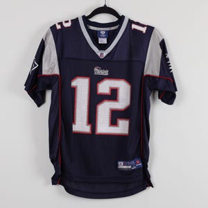 May include: A navy blue short-sleeve sports jersey featuring the number 12 in white with red outlines on the front and shoulders. The jersey includes silver accents on the sleeves, a V-neck collar, and the Patriots logo printed on the chest.