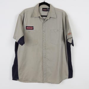 May include: A tan short-sleeved button-up shirt with navy blue side panels. The shirt has two chest pockets and a collar. The Dickies logo is on the left chest and sleeve. The shirt is hanging on a black hanger.