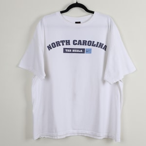 May include: White t-shirt featuring the text "NORTH CAROLINA TAR HEELS" in navy blue arched lettering. The Nike logo is on the right side of the text. The shirt is hanging on a black hanger.