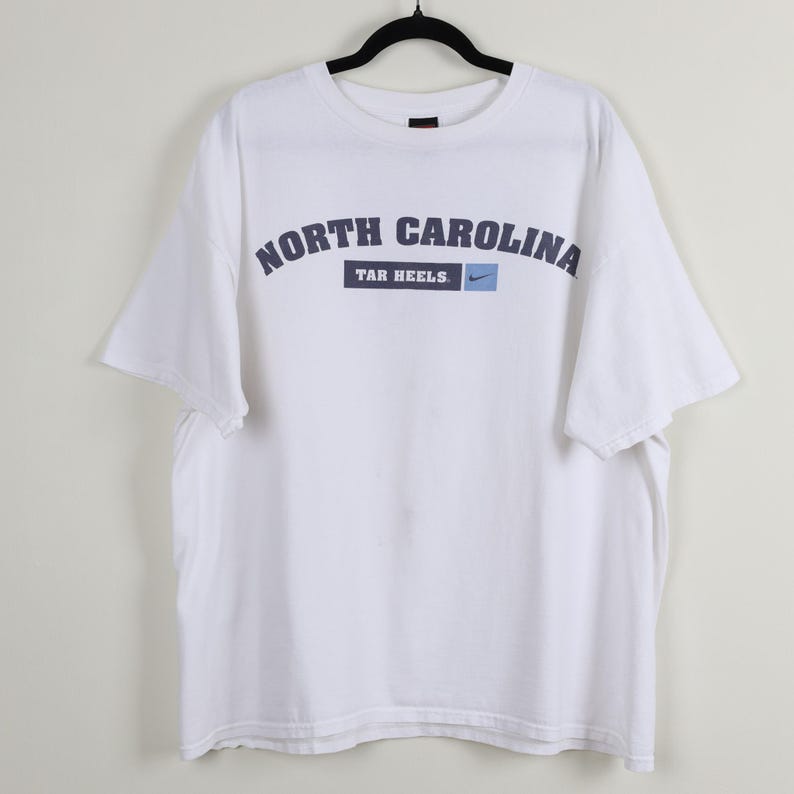 May include: White t-shirt featuring the text "NORTH CAROLINA TAR HEELS" in navy blue arched lettering. The Nike logo is on the right side of the text. The shirt is hanging on a black hanger.