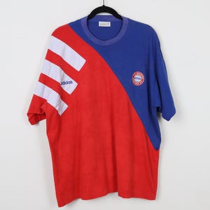 May include: A red, white, and blue short-sleeved t-shirt with the Adidas logo and the FC Bayern Munich logo. The shirt has a diagonal color block design.