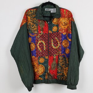 May include: A long-sleeve zip-up jacket featuring a patchwork design of red, blue, and dark green panels. The fabric displays a repeating pattern of gold-colored chains, tassels, and horseshoe motifs. The sleeves and collar are solid dark green.