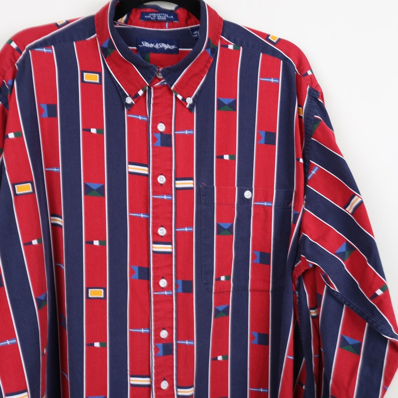 Puede incluir: Una camisa de manga larga con botones a rayas rojas y azules con un patr&oacute;n geom&eacute;trico. La camisa tiene un bolsillo en el lado izquierdo y un cuello abotonado.