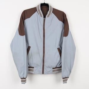 May include: A light blue and brown bomber jacket with a full zipper closure. The jacket features brown shoulder panels, a brown collar, and brown trim on the cuffs and waist. The jacket is hanging on a black hanger.
