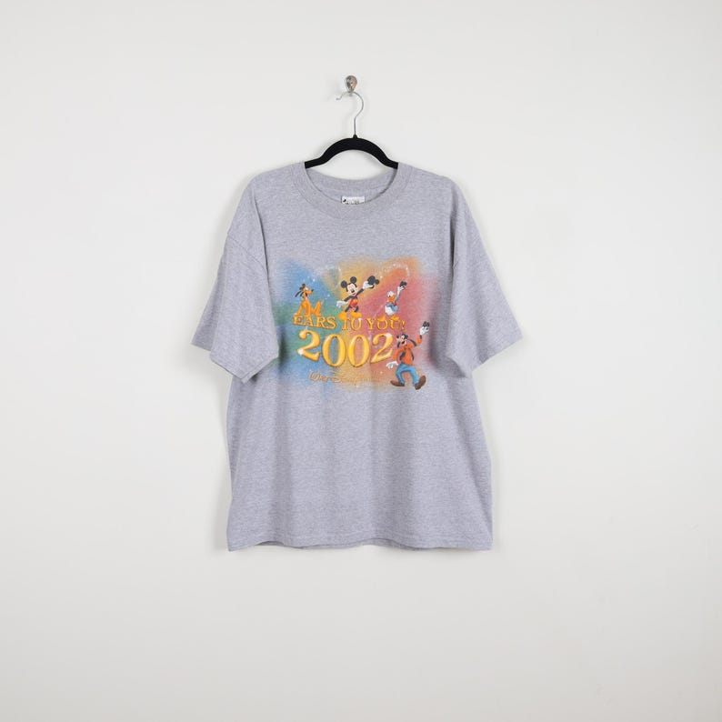 Pode incluir: Uma camiseta cinza com um gr&aacute;fico colorido de personagens da Disney, incluindo Mickey Mouse e Pateta. O design inclui o texto "EARS TO YOU" e "2002". A camiseta tem mangas curtas e gola redonda.