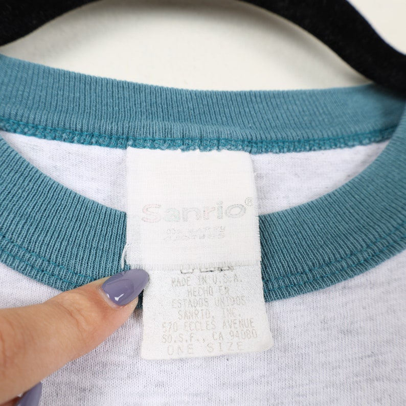 Puede incluir: Camiseta blanca con cuello de canal&eacute; azul turquesa y una etiqueta blanca de Sanrio. La etiqueta dice "Sanrio" y "Made in U.S.A. Hecho en Estados Unidos Sanrio, Inc. 570 Eccles Avenue So.S.F., CA 94080 One Size"