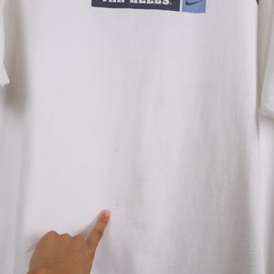 May include: White t-shirt featuring the text "AIR HEELS" in a navy blue rectangle, with a blue Nike swoosh logo. The shirt is made of a soft material, possibly cotton.