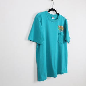 Puede incluir: Camiseta turquesa con un bolsillo en el pecho izquierdo. La camiseta tiene un gr&aacute;fico colorido en el bolsillo que dice "Aron's Surf Shop". La camiseta est&aacute; colgada en una percha negra contra una pared blanca.