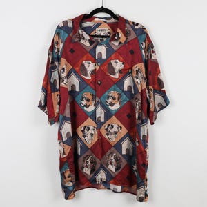 May include: Short-sleeved button-down shirt with a repeating pattern of dog faces and dog houses. The shirt features a color palette of burgundy, teal, tan, and navy blue. The shirt is hanging on a black hanger.