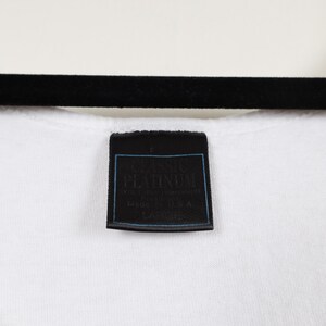 May include: Close-up of a white t-shirt with a black label. The label has the word "PLATINUM" in white text with a blue border. The shirt is hanging on a black hanger.