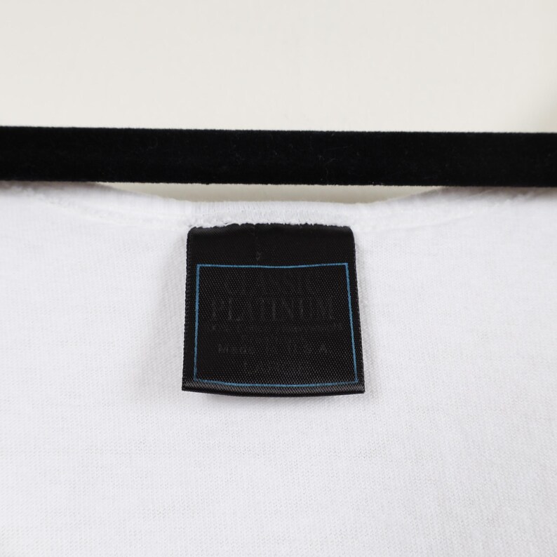 May include: Close-up of a white t-shirt with a black label. The label has the word "PLATINUM" in white text with a blue border. The shirt is hanging on a black hanger.