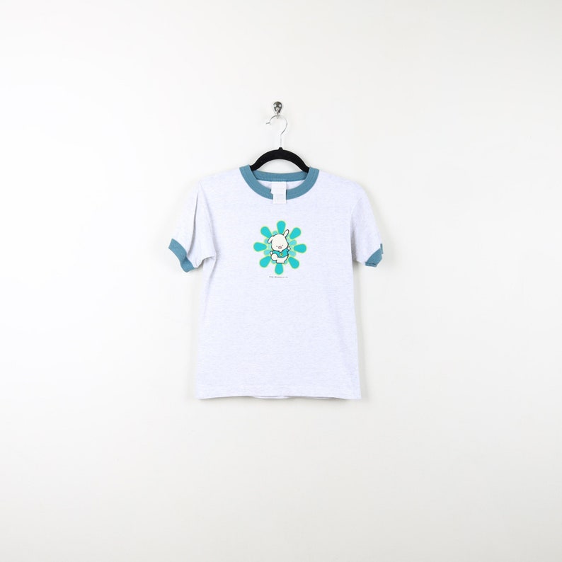 Puede incluir: Una camiseta blanca con un ribete azul. La camiseta presenta un gr&aacute;fico de un conejito blanco en un dise&ntilde;o de flor azul y verde. El texto "The Original" est&aacute; impreso debajo del gr&aacute;fico.