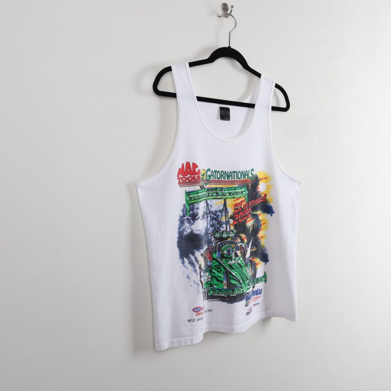 May include: White tank top with a colorful graphic print featuring the text "MAC TOOLS GATORNATIONALS". The graphic includes a race car and palm trees. The tank top is hanging on a black hanger against a white wall.