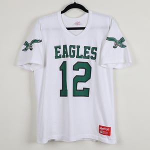 May include: White v-neck jersey with the word "EAGLES" in green block letters and the number "12" in green. Green eagle logos are on the sleeves. A red and white Rawlings tag is at the bottom.