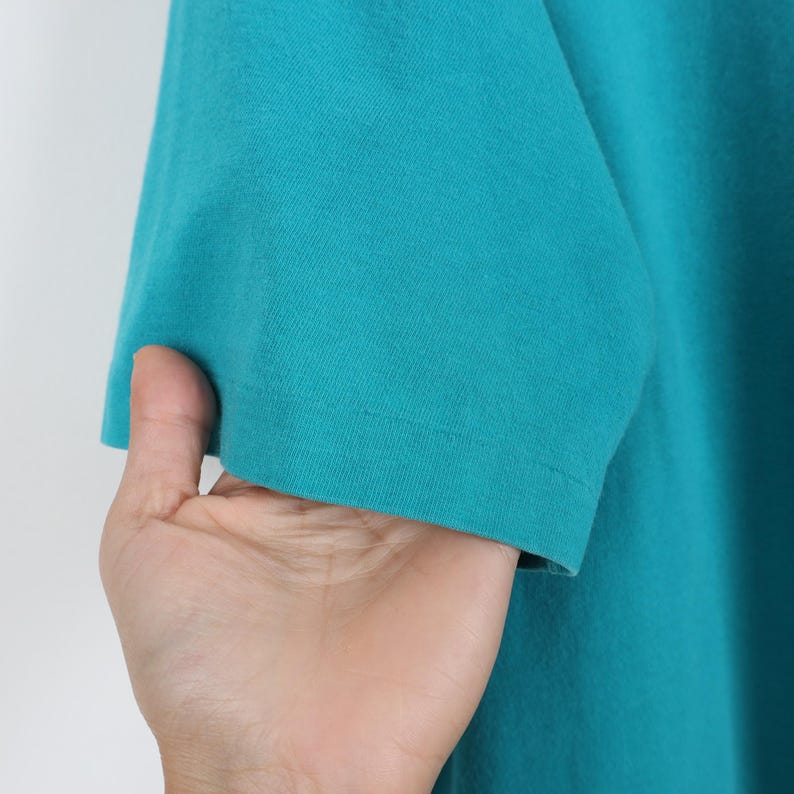 Puede incluir: Primer plano de una camiseta turquesa de manga corta. Se ve la manga, con un borde con dobladillo. La tela parece ser un material de punto suave. La camiseta es de color liso, sin patrones ni gr&aacute;ficos visibles.