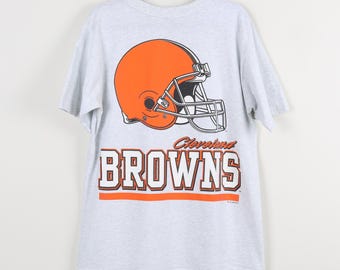May include: A light gray t-shirt featuring an orange football helmet graphic. Below the helmet, the word "BROWNS" is printed in bold, white letters with an orange outline. The shirt is hanging on a black hanger.